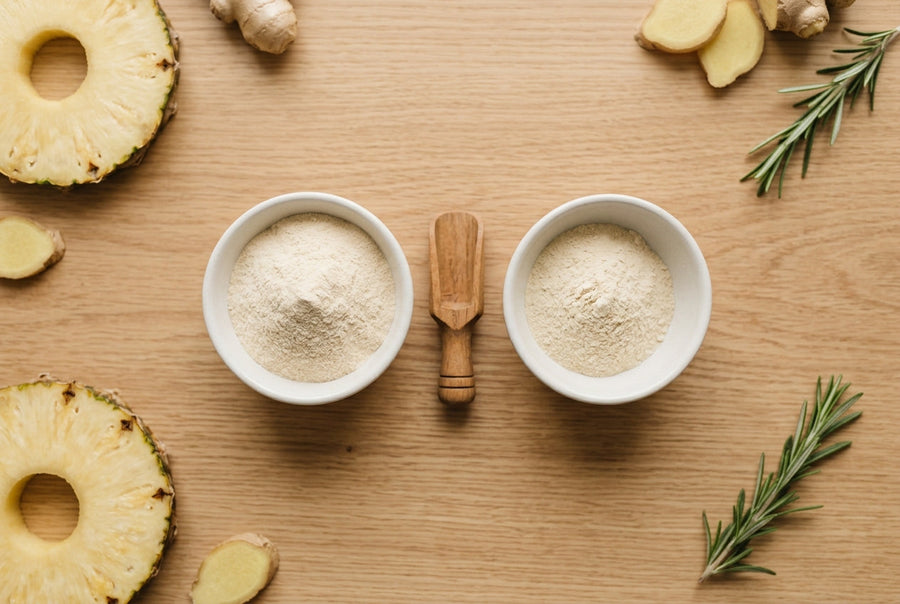 Two bowls of pale collagen powder with a wooden scoop, pineapple, ginger, and rosemary on a light oak table.