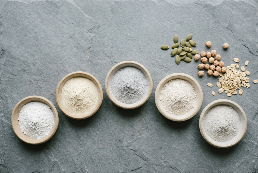 Five small ceramic dishes of white mineral powders on grey slate beside pumpkin seeds, chickpeas, and rolled oats.