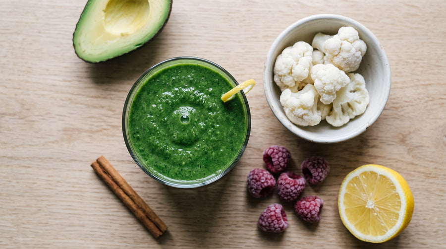 Green smoothie in a glass beside frozen cauliflower, a quarter avocado, a few raspberries, lemon, and a cinnamon stick.