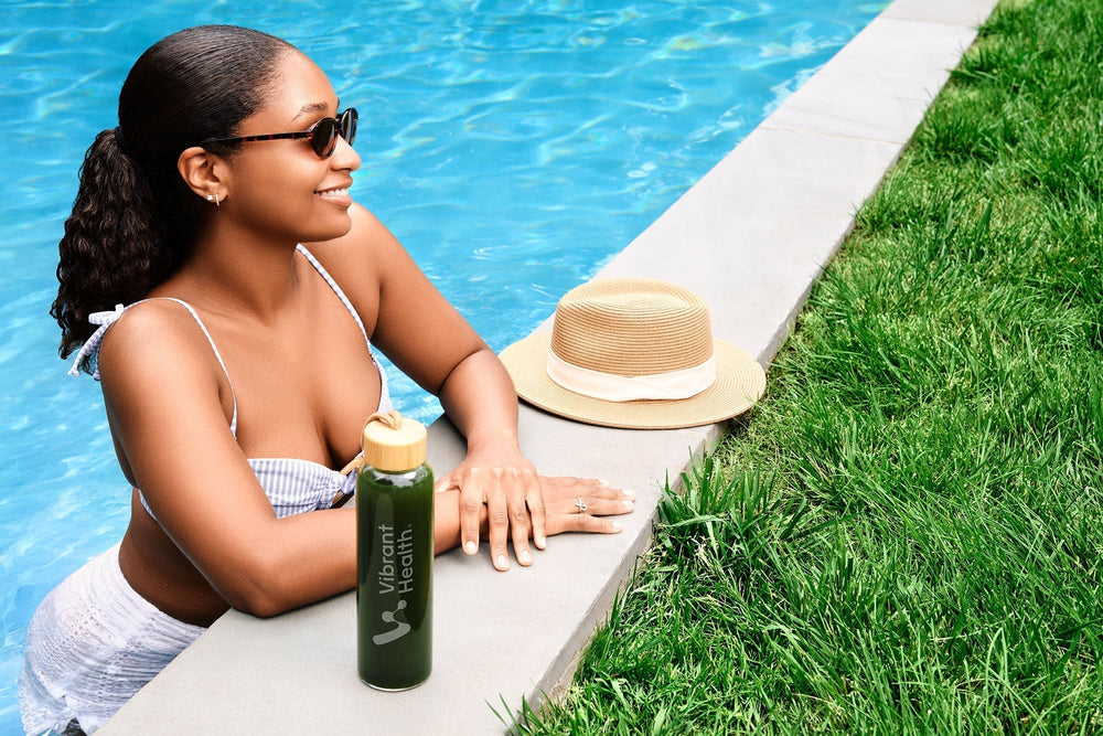 A young women in a swimming pool with a super greens drink and a straw hat next to her.