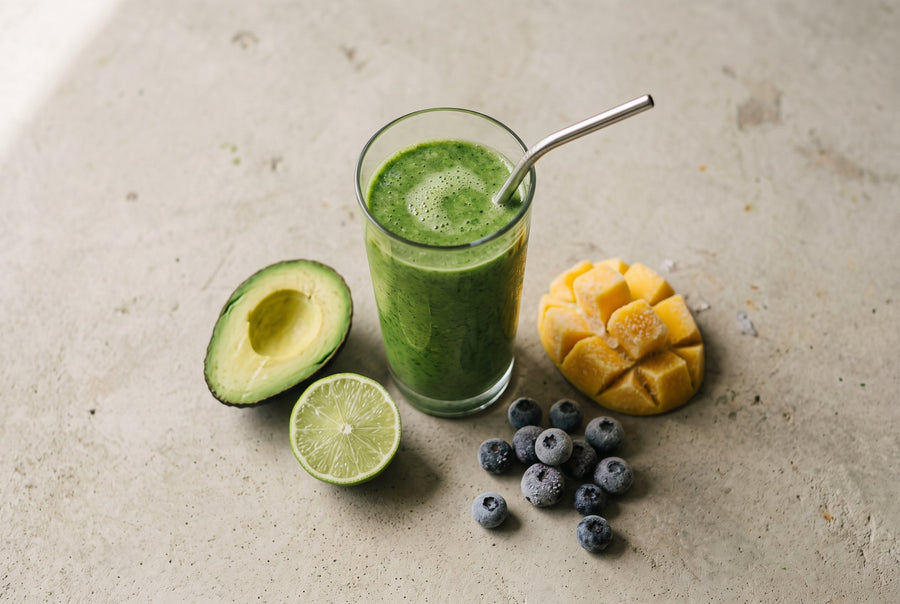 Overhead view of a green smoothie in a tall glass beside frozen mango, blueberries, halved lime, and avocado.