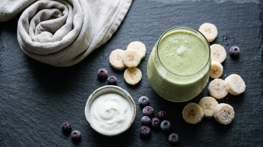 Overhead view of a pale green shake in a glass beside frozen banana slices, yogurt, blueberries, and a folded towel.