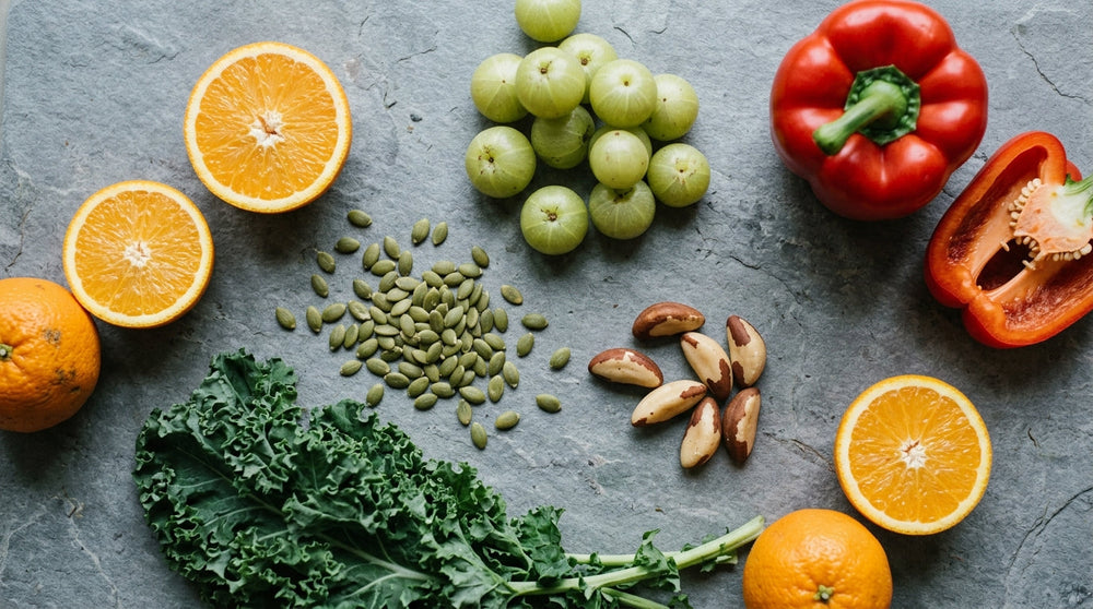 "Overhead shot of whole foods scattered on a matte grey stone surface, including halved oranges, fresh kale, pumpkin seeds, Brazil nuts, red bell pepper, and amla berries.