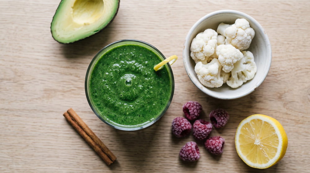 Green smoothie in a glass beside frozen cauliflower, a quarter avocado, a few raspberries, lemon, and a cinnamon stick.