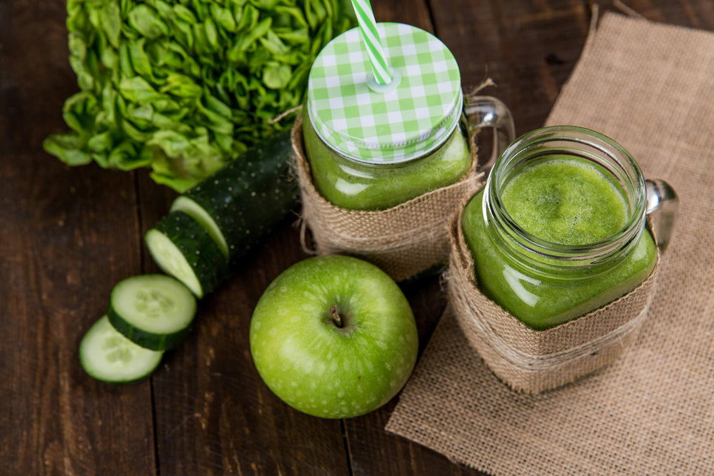 2 cups of Greens powder shakes on a wooden table with a green apple and some other vegetables on the side. 