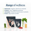 Two stand-up pouches of VITALITY MAXIMUM VIBRANCE plant-based protein powder are displayed on a white reflective surface with wellness-themed props including a sprig of green grass, a small glass vase of parsley, a jar of yellow grains, and a bowl of red gummies. Text on the image lists product benefits including multivitamin, probiotics, antioxidants, and protein.